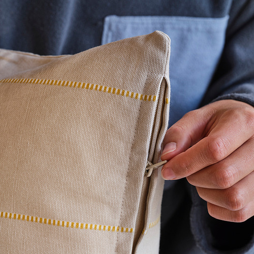 Primer plano de una mano sosteniendo una funda de cojín de algodón beige con motivos geométricos amarillos. La cremallera está parcialmente abierta, lo que demuestra su facilidad de uso.