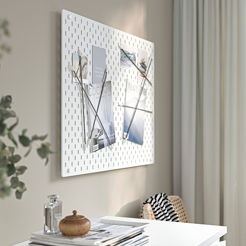 Un tablero perforado SKÅDIS blanco montado en la pared muestra fotos y revistas organizadas. Un elegante escritorio debajo sostiene una planta decorativa, un frasco y una bolsa tejida.