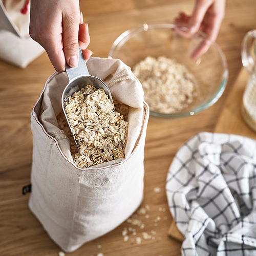 Persona sacando avena de una bolsa de tela blanca sobre una mesa de madera. Otros artículos de cocina cercanos.