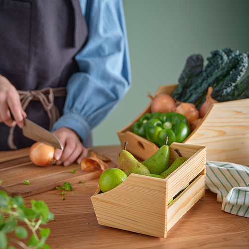 Una persona está cortando cebollas en una tabla de cortar de madera, con otras verduras en cajas de madera sobre una mesa.