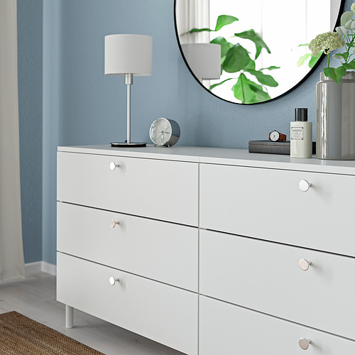 Tocador blanco con pomos de cajón, lámpara plateada, reloj, espejo, plantas verdes, pared azul.