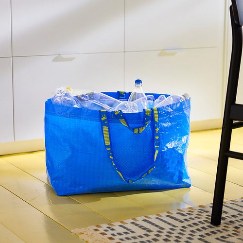 Bolsa de compras reutilizable azul, llena de botellas de plástico, colocada sobre un suelo de baldosas junto a una silla negra y una alfombra con motivos geométricos.