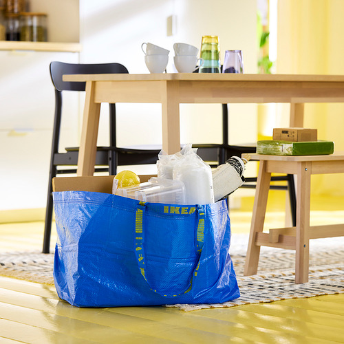 Bolsa azul de ikea FRAKTA, llena de cajas y artículos, colocada sobre un suelo de madera junto a una mesa de madera con sillas.