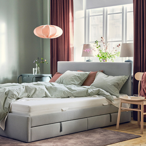 Un acogedor dormitorio con un gran colchón ÅBYGDA gris sobre una estructura de cama junto a una mesa auxiliar de madera con una lámpara y flores.