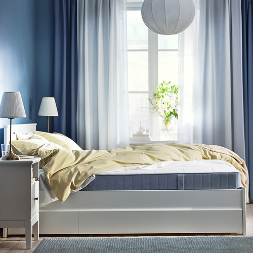 Acogedora habitación con cortinas azules, cama blanca, manta amarilla, lámparas y planta.