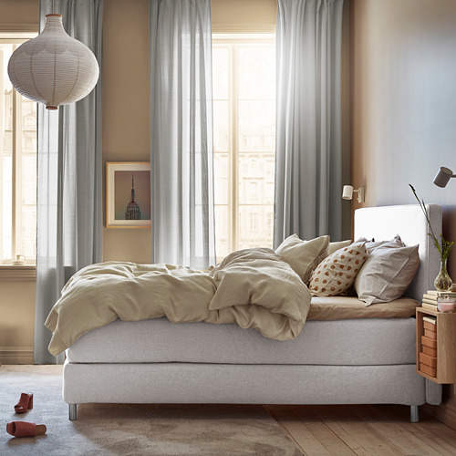 Dormitorio con ropa de cama beige, cama blanca, grandes ventanales con cortinas, arte de pared, mesita de noche con libros y lámpara, gran lámpara blanca colgante.