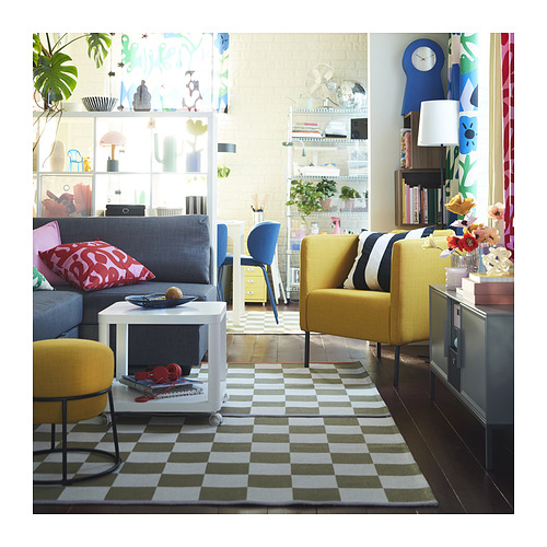 Sala de estar luminosa con sofá gris, sillones amarillos, alfombra a cuadros, mesa de centro blanca, decoración colorida y gran ventanal.