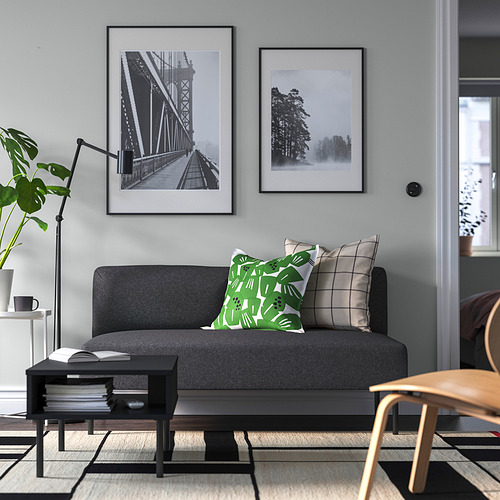 Salón moderno con sofá LILLEHEM gris, almohadas verdes, mesa de centro negra, silla de madera, alfombra, lámpara, libros, arte.