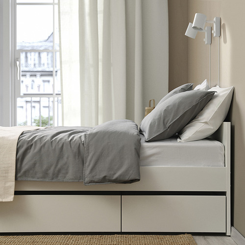 Estructura de cama con dos cajas de almacenamiento debajo de la cama, visibles a través de un cajón parcialmente abierto, con lámpara de pared encima.