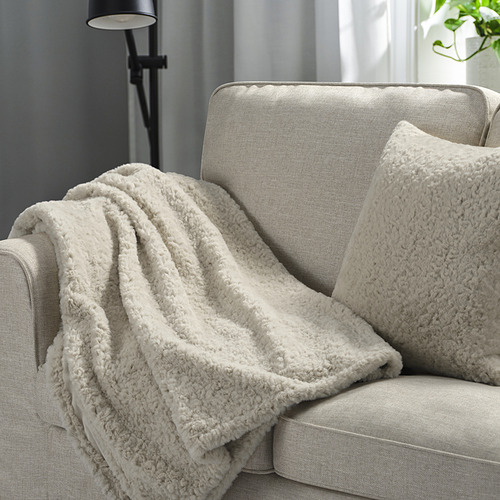 Manta y almohada NÄTNÄVA beige sobre sofá gris, creando un ambiente acogedor.