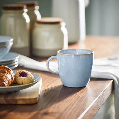 Una taza azul claro de FÄRGKLAR se encuentra sobre una mesa de madera cerca de pasteles y platos, mostrando su diseño simple y funcional en un ambiente acogedor.