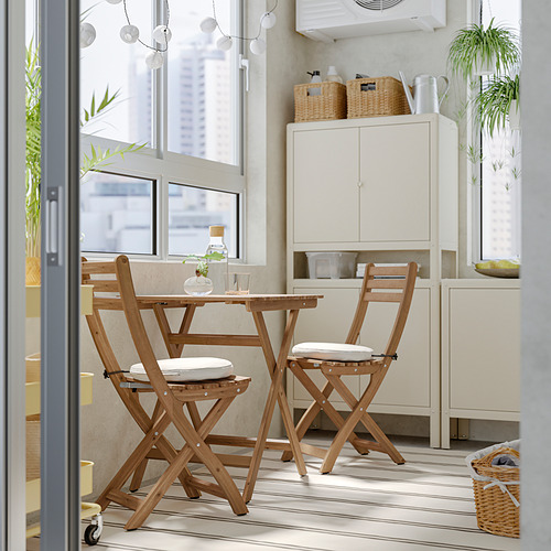 Juego de comedor de madera con cojines en un espacio acogedor. Mesa de acacia y sillas plegables, cojines blancos, cerca de la ventana con vegetación.