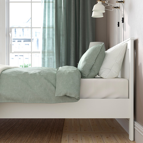 Dormitorio luminoso con estructura de cama SONGES blanca, almohadas de color verde claro y suelo de madera. La cama tiene lados ajustables y patas altas.