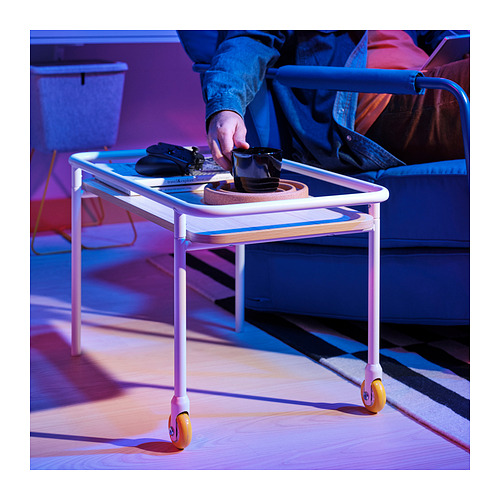 Mesa auxiliar blanca con ruedas naranjas, bandeja de sujeción, teléfono y taza de café. La mano de la persona alcanza la taza. Ideal para bocadillos y configuración de juegos.
