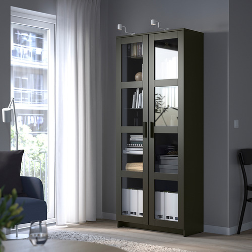 Librería BRIMNES con puertas de vidrio cerca de la ventana, con libros y decoración, silla azul al lado.