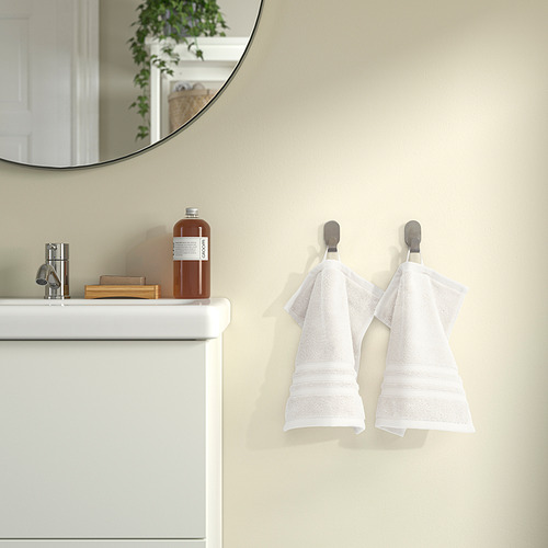 Toalla BROKGLIM blanca en gancho de pared en baño, felpa y grande, cerca de lavabo con espejo y botella.