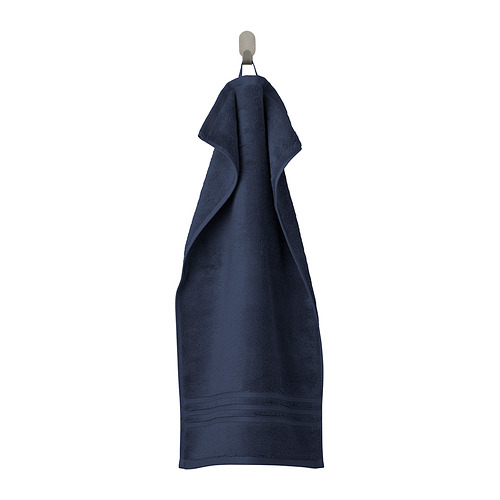 Una toalla BROKGLIM azul oscuro cuelga cuidadosamente de un gancho. Confeccionado en algodón orgánico suave y absorbente con un borde tejido.