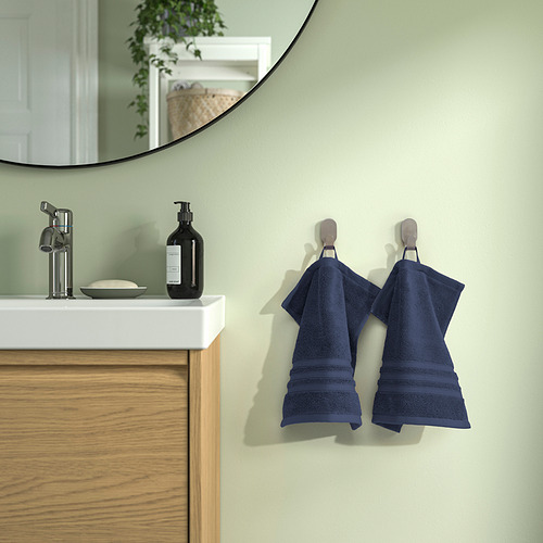 Toallas BROKGLIM azules en ganchos cerca de lavabo moderno con espejo. Algodón orgánico, borde elegante. Paredes verdes, armario de madera. Diseño limpio y contemporáneo.