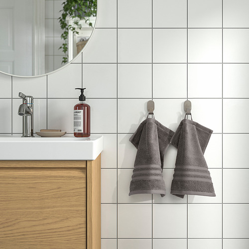 Dos toallas BROKGLIM grises cuelgan cuidadosamente en ganchos junto a un lavabo blanco con un armario de madera.