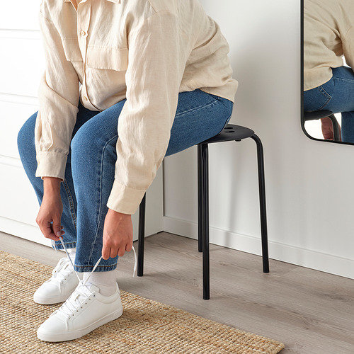 Persona sentada en un taburete MARIUS negro, atando una zapatilla blanca, ambiente informal.
