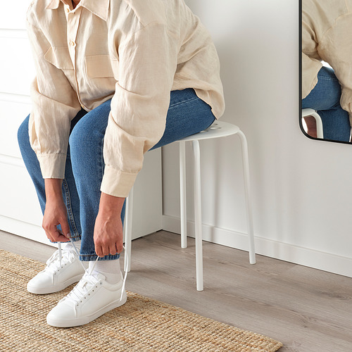 Persona atando zapatillas sobre taburete blanco. Habitación minimalista con espejo, alfombra.