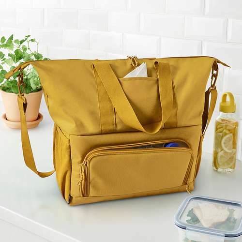 Gran bolsa de mano aislante KLOTPIL de color mostaza sobre la mesa, rodeada de una planta, una botella de agua y una lonchera.