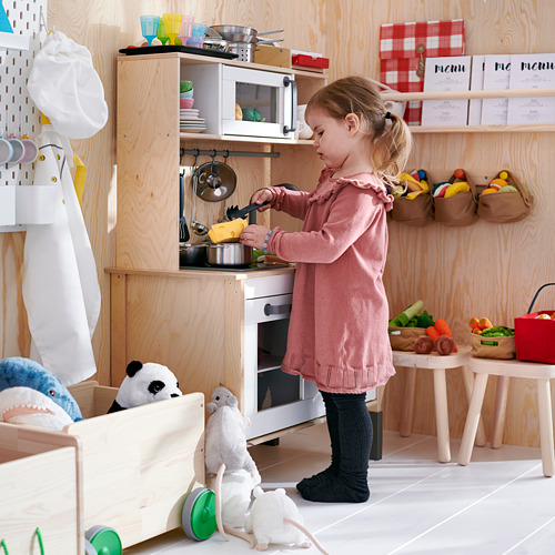 La niña juega en la cocina de madera, con animales de peluche, tazas de colores y delantal colgante, preparando la comida con un juego de juguetes.