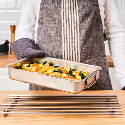 Una persona con un delantal sostiene un molde para hornear verduras asadas sobre una mesa con rejillas metálicas para asar.
