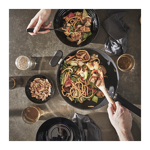 Dos personas cocinando fideos en wok negro; uno revuelve, otro sirve.
