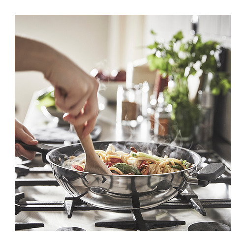 La persona revuelve la comida en un wok MIDDAGSMAT de acero inoxidable en una estufa de gas, rodeada de hierbas y utensilios de cocina.