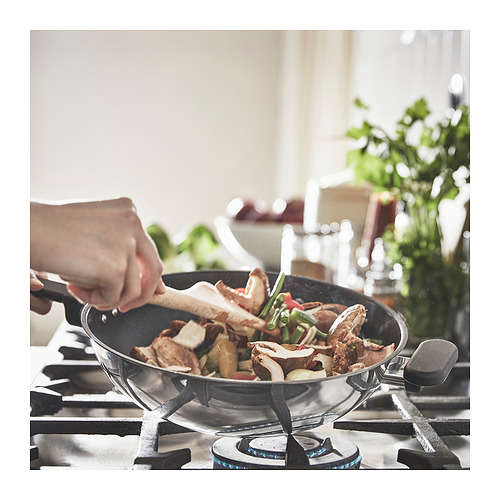 Persona cocinando en un wok de vidrio transparente en una estufa. Ingredientes visibles en el interior. Ambientación de la cocina con plantas verdes.
