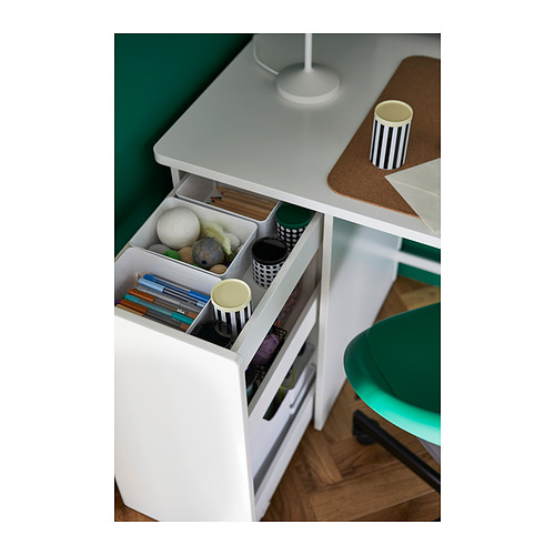 Escritorio blanco con cajón abierto que muestra bolígrafos, libros y juguetes pequeños. Pared verde y silla a juego cerca.