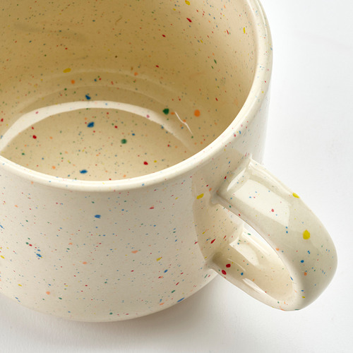 Taza de cerámica con motas multicolores, asa resistente, colocada sobre superficie blanca.