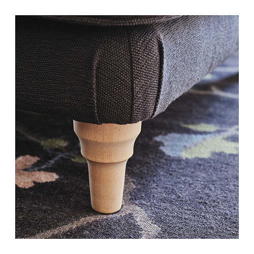 Primer plano de una pata de madera con vetas visibles, sosteniendo una pieza de tela oscura sobre una alfombra estampada.