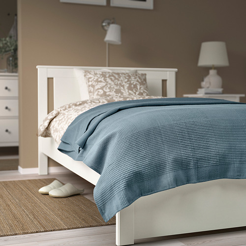 Colcha de algodón azul sobre una gran cama blanca, con almohadas, lámpara y zapatillas cerca.