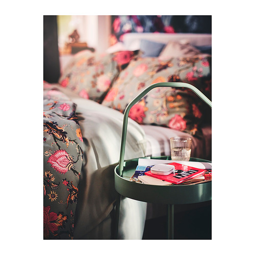Pequeña mesa auxiliar verde junto a la cama, con libros, control remoto y vidrio.