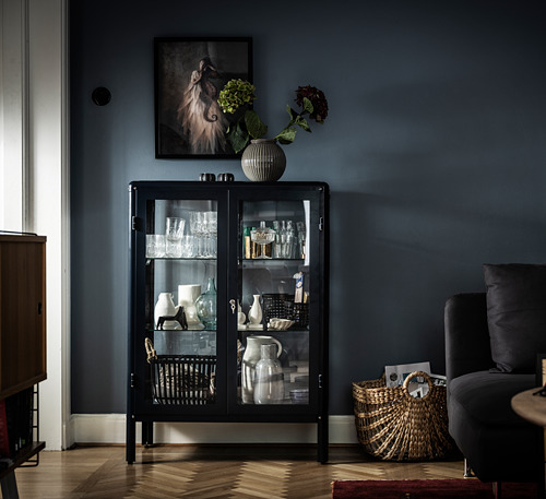 Mueble FABRIKÖR negro con puertas de cristal, con jarrones y vasos, rematado con un jarrón a rayas con flores. Decoración moderna y elegante de la sala de estar.