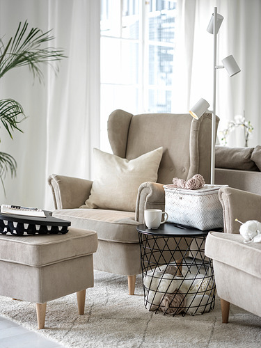 Sala de estar moderna con sillón orejero STRANDMON beige, otomana a rayas, mesa auxiliar de metal y plantas.