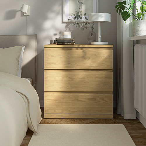 Un cofre de madera clara, más pequeño que la cama, con una lámpara, plantas y libros encima.