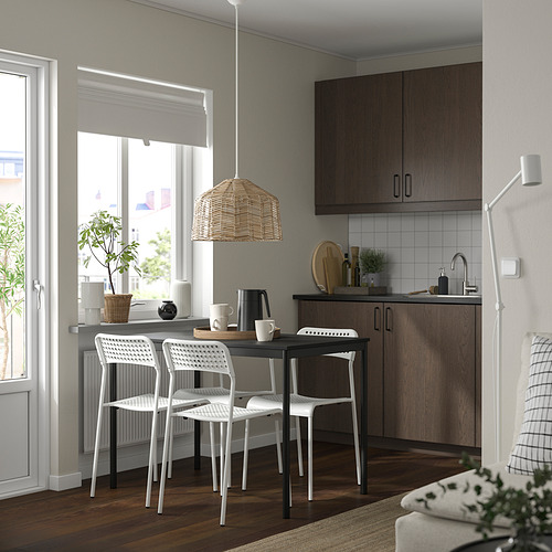 Acogedora cocina con sillas apilables sansberg blancas, mesa de madera y lámpara colgante de mimbre. Armarios de madera oscura y gran ventanal con plantas.