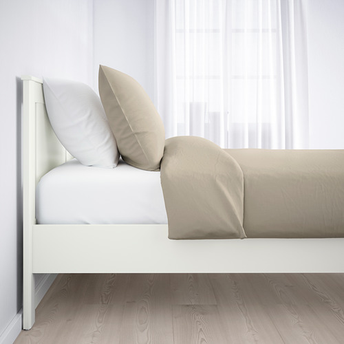 Cama blanca minimalista con almohadas beige, sobre cortinas blancas transparentes sobre un suelo de madera.