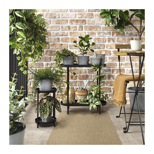 Esta imagen muestra un acogedor entorno exterior con una alfombra LÄTTBETONG beige y marrón, varias plantas de interior y una pared de ladrillos.