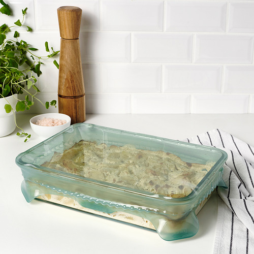 Fuente para hornear de vidrio con tinte verde, que sostiene alimentos horneados, colocados sobre un paño a rayas, junto a la planta, un tazón de sal y un molinillo de pimienta.