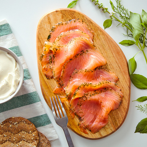 Plato de madera con salmón curado en rodajas de naranja, guarnición de eneldo, tenedor, pan y salsa de crema.