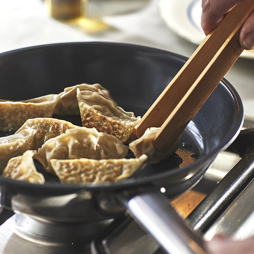 Una mano usa palillos para recoger albóndigas de una sartén negra en una estufa, enfatizando la superficie antiadherente de las sartenes y la facilidad de cocción.