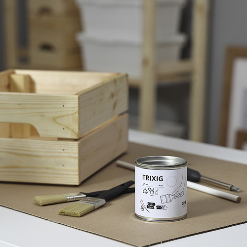 Una pequeña lata blanca de pintura TRIXIG descansa sobre un cartón, flanqueada por un pincel y un destornillador. Cerca hay cajas de madera, lo que sugiere proyectos de bricolaje.