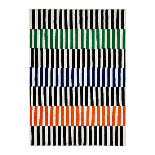 Una alfombra rectangular VÄNDPLATS de tom hedqvist, con rayas negras, verdes, azules y naranjas, hecha de polipropileno.