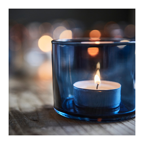 Una vela encendida en un soporte de vidrio azul se encuentra sobre una superficie de madera, emitiendo un cálido resplandor. El soporte tiene un diseño elegante y moderno con un acabado liso.