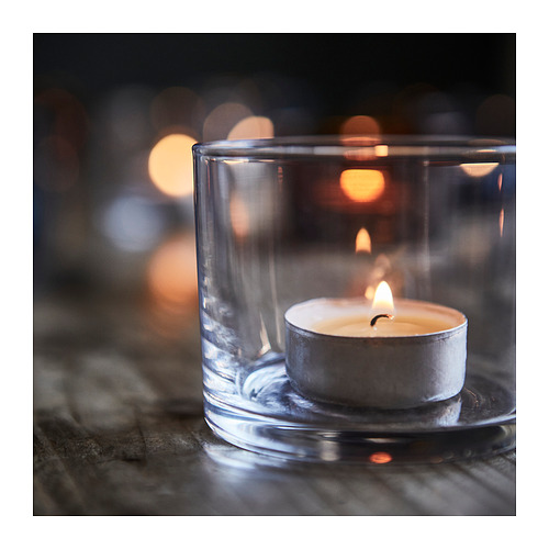 Vela pequeña en soporte de vidrio transparente; mecha iluminada, brillo suave, superficie reflectante.