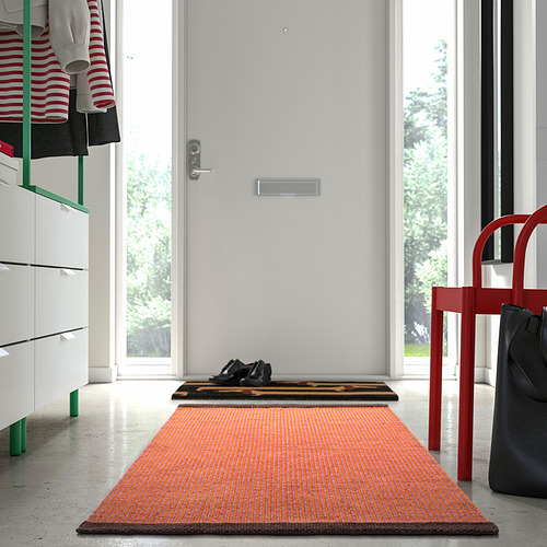 Alfombra VÄGOMRÅDE naranja y marrón en el pasillo, cerca de la puerta blanca y la silla roja.